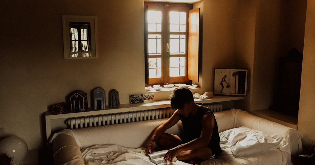 Iemand zit voorovergebogen op bed in een kamer met een open raam